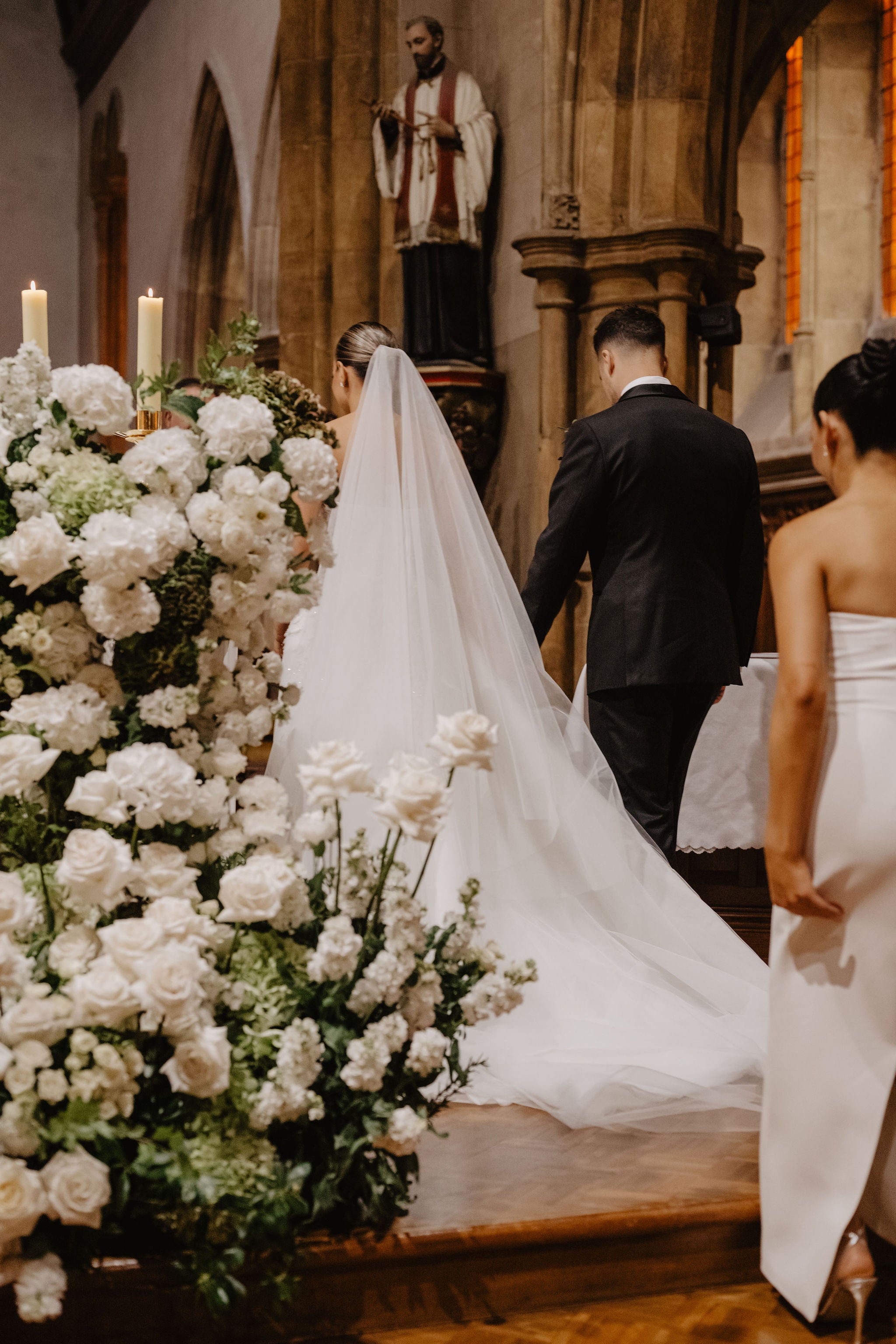 White roses, hydrangea and stock banked in a tall arrangement with two lit candles beside it, inside a stone church, as a bride with a long veil and a groom walk away up the step to the altar.